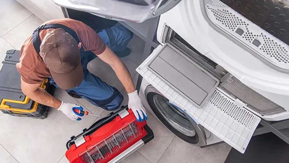 Een wasdroger wordt gerepareerd, met een rode gereedschapskist en een grijze aan de kant. De droger heeft een open filter.