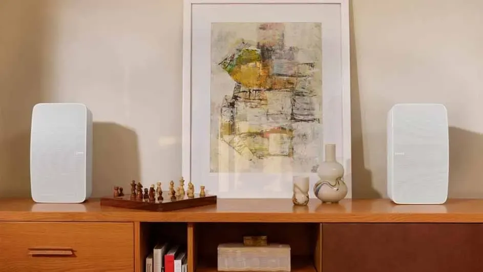 Woonkamerinterieur met houten dressoir tegen een beige muur. Er staan twee witte luidsprekers, een schilderij, een schaakbord en vazen op het dressoir.