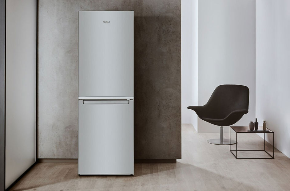 Frigo gris argenté dans une pièce moderne. Une chaise sombre et une table avec des bouteilles sont visibles au fond.