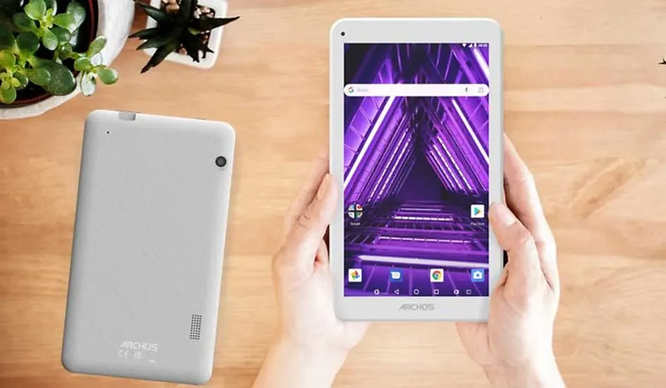 Tablette blanche avec écran violet dans les mains, à côté d'une autre tablette et de plantes sur une table en bois.