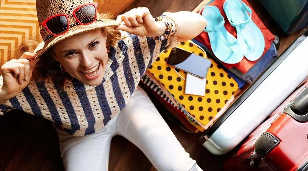 Valise ouverte avec vêtements, smartphone et sandales bleues. Chapeau de paille avec lunettes de soleil rouges sur le dessus.