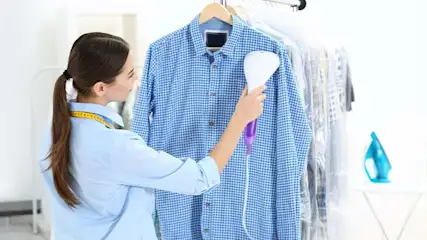 Une chemise bleue à carreaux est suspendue à un cintre. Un défroisseur blanc et violet est utilisé sur la chemise.