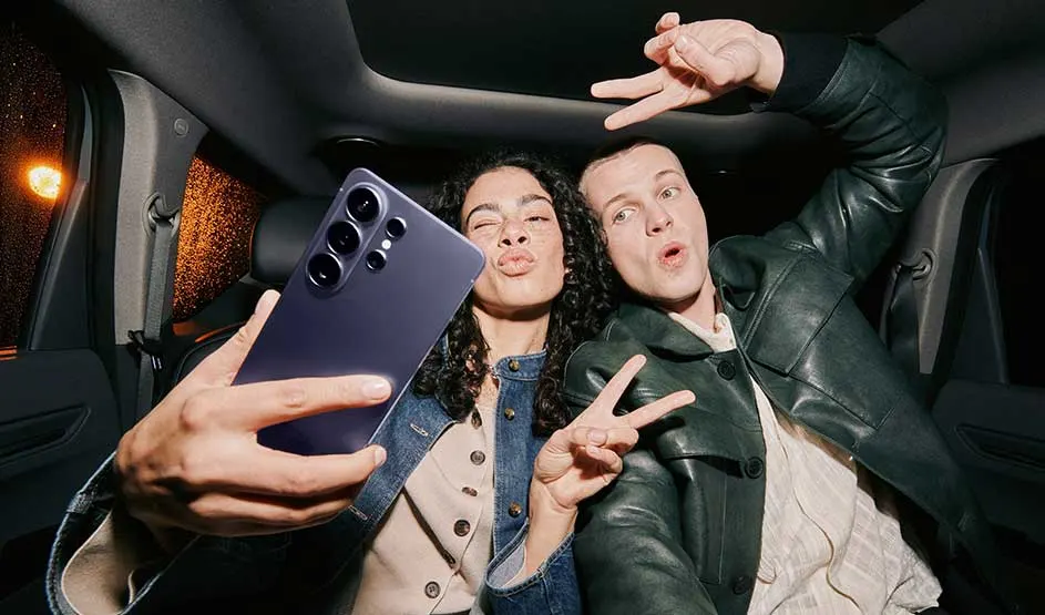 Twee vrienden maken een speelse selfie op de achterbank, houden een paarse Samsung-telefoon voor de lens en vormen vrolijk victorygebaren.