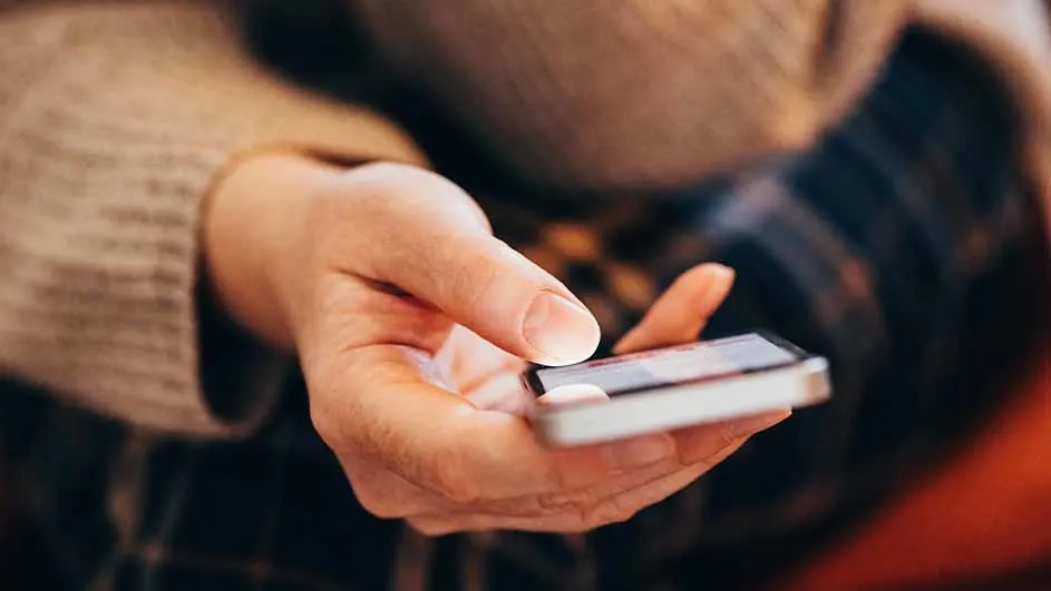 Een hand bedient een witte smartphone met een helder scherm, gekleed in een warme trui, zittend op een geruite broek.