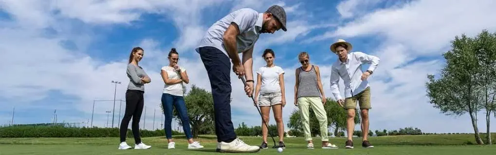 Un groupe regarde un homme jouer au golf sur un terrain vert avec des arbres et un ciel bleu nuageux en arrière-plan.