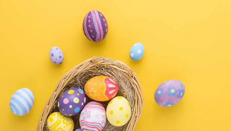 Des œufs de Pâques décorés dans un panier en osier sur un fond jaune, certains œufs sont éparpillés autour du panier.