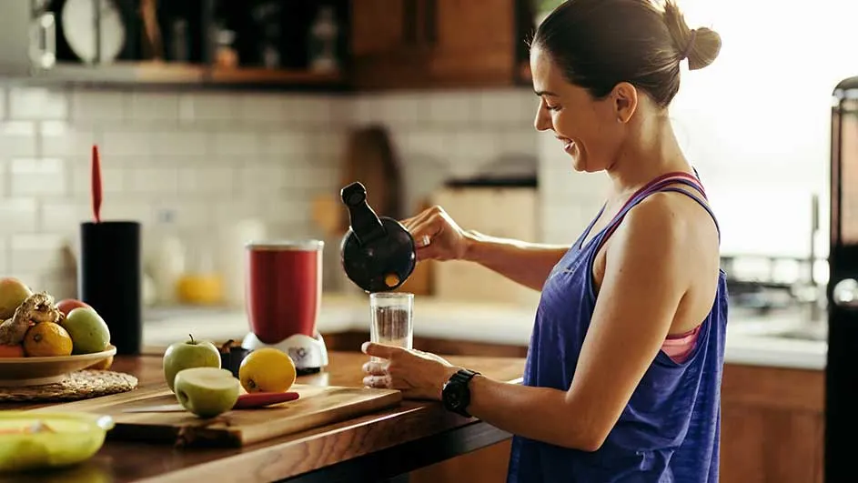 Op een houten aanrecht staat een blender naast een snijplank met fruit. Iemand giet vloeistof uit een kan in een glas.