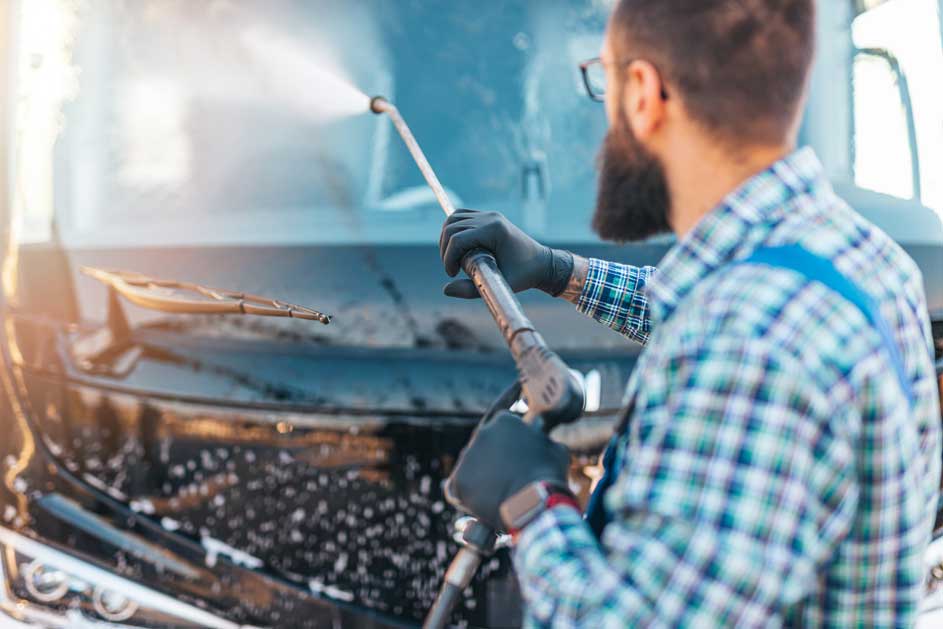 Een blauwe vrachtwagen wordt gewassen met een hogedrukreiniger. Schuim bedekt de voorkant. De ruitenwisser is zichtbaar.