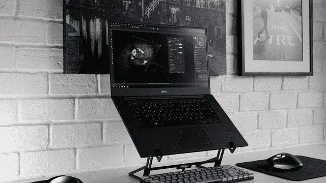 Een zwart-wit foto toont een laptop op een standaard, met een extern toetsenbord en muis. De achtergrond is een bakstenen muur met lijsten.