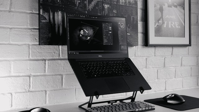 Un ordinateur portable noir sur un support, un clavier et une souris sur un bureau blanc, devant un mur de briques blanches. Tableaux au mur.