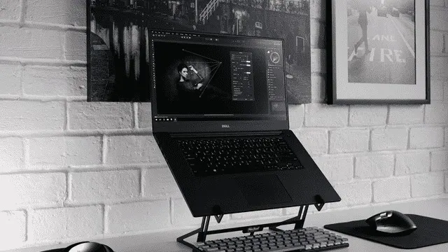 Un ordinateur portable noir sur un support, un clavier et une souris sur un bureau blanc, devant un mur de briques blanches. Tableaux au mur.