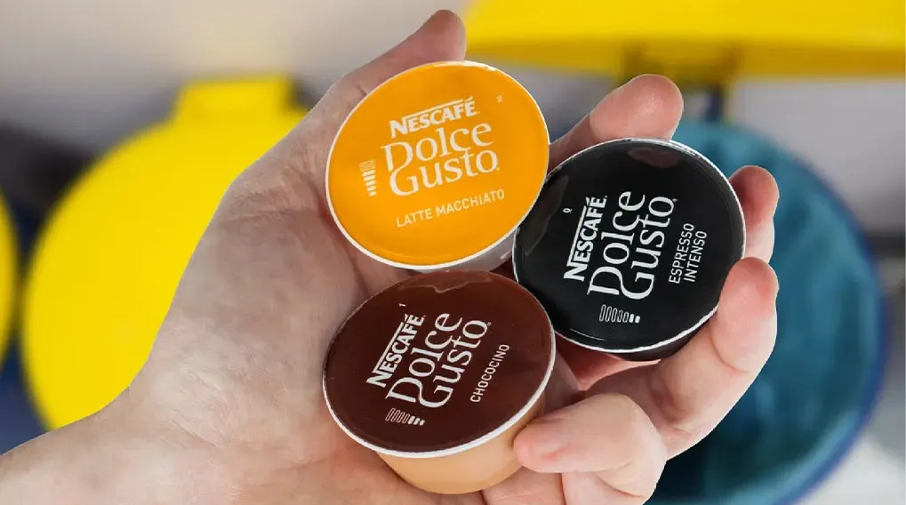 Een hand houdt drie Nescafé Dolce Gusto koffiecapsules vast: oranje 'Latte Macchiato', bruin 'Chococino' en zwart 'Espresso Intenso'.