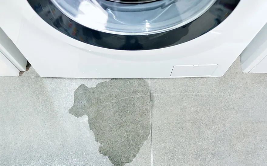 Vue rapprochée d'un lave-linge blanc sur un sol carrelé gris avec une flaque d'eau.