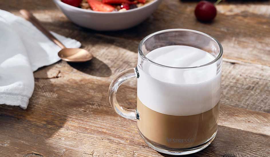 Een glazen mok met koffie en melk staat op een houten tafel. Een kom fruit en een lepel zijn ook zichtbaar.