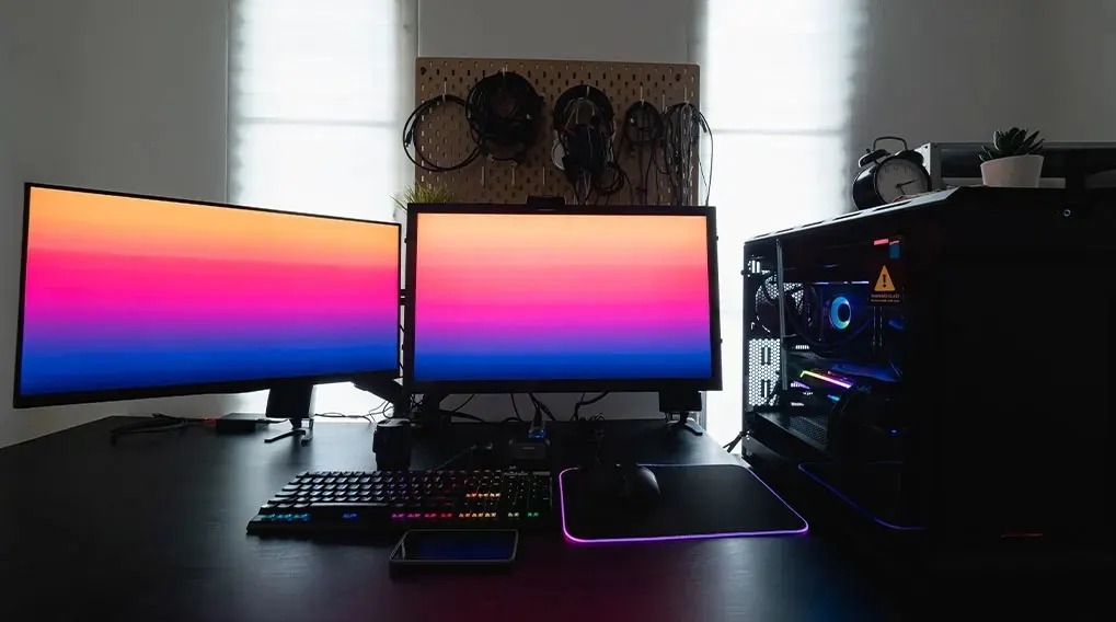 Bureau noir avec deux écrans affichant un dégradé de couleurs, un clavier, une souris et une unité centrale d'ordinateur noire.