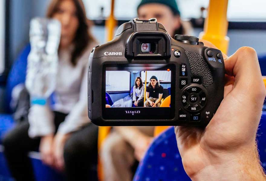 Een zwarte Canon camera wordt vastgehouden, met een scherm dat een foto toont van mensen in een bus.