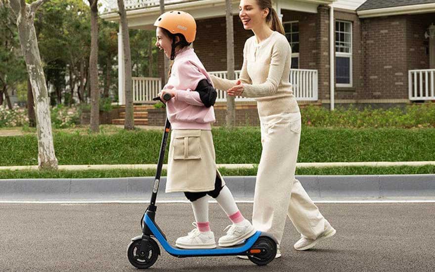 Een meisje met helm rijdt lachend op een elektrische step, terwijl haar moeder achter haar duwt op een rustige woonstraat.