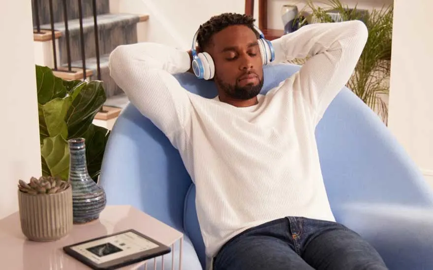 Iemand zit in een blauwe stoel met een witte trui en een koptelefoon. Er staat een e-reader en een vaas op tafel.