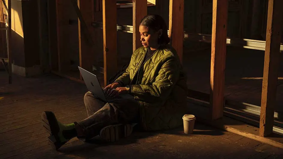 Een laptop op een houten vloer naast een kartonnen beker, omringd door houten balken in een kamer met weinig licht.
