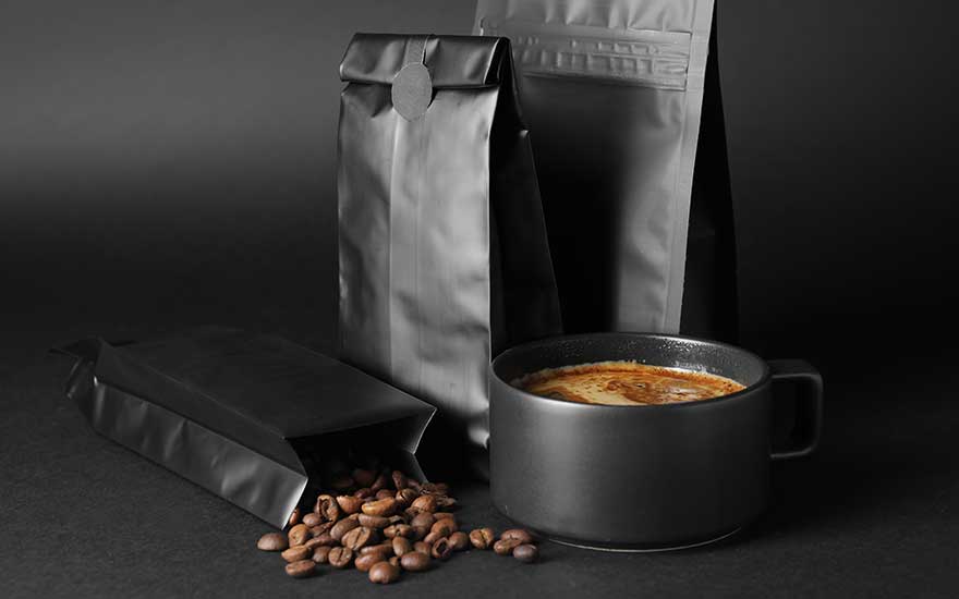 Trois paquets noirs, grains de café et tasse noire remplie de café sur fond noir.