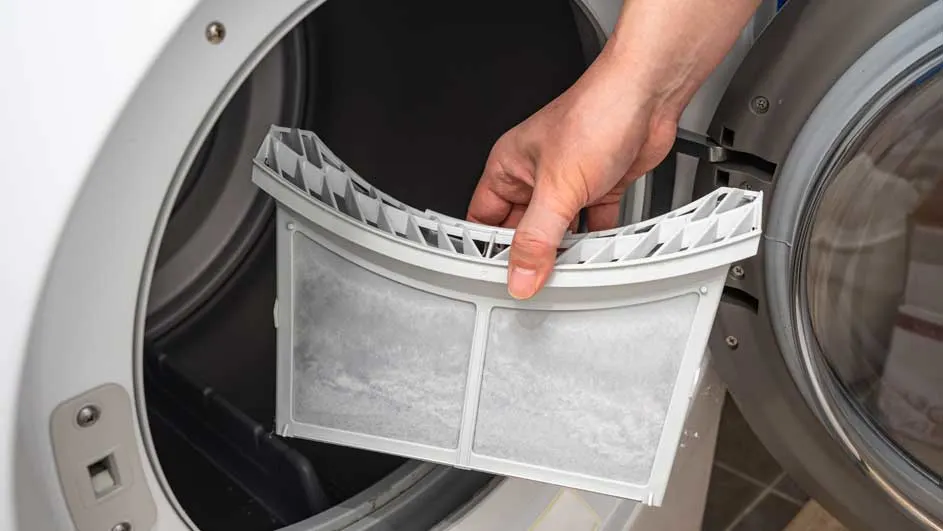 Une main retire un filtre à charpie gris d'un sèche-linge blanc. L'intérieur du sèche-linge est sombre.