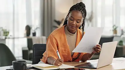 Une femme travaille à un bureau avec un ordinateur portable, des papiers, un carnet et un stylo. Une lampe est sur le bureau.