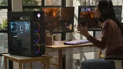 Un écran affiche un jeu vidéo. Un ordinateur de bureau bleu est à côté. Un clavier et une souris sont sur une table en bois.