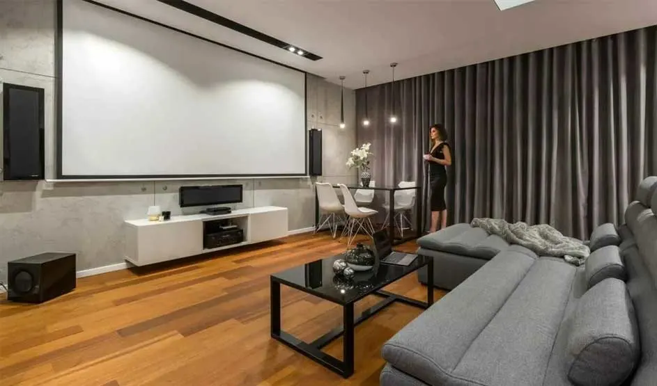 Een moderne woonkamer heeft een groot projectiescherm, grijze bank en warme houten vloer.