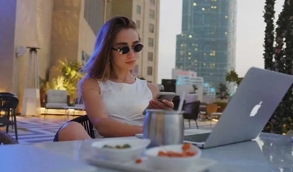 A woman in sunglasses works at a rooftop café, checking her phone beside an open Apple MacBook, with snacks and city towers in the dusk.