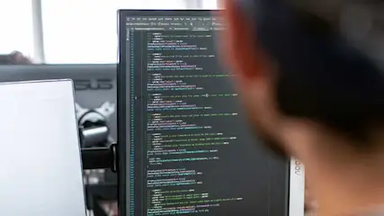 Een computerscherm toont programmeercode met verschillende kleuren tekst op een donkere achtergrond. Een ander scherm is gedeeltelijk zichtbaar links.