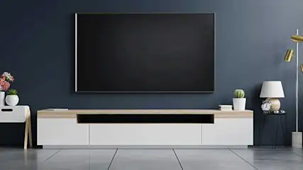 Un écran noir est fixé au mur bleu au-dessus d'une unité multimédia blanche avec un dessus couleur bois clair. Des plantes et des lampes sont visibles.