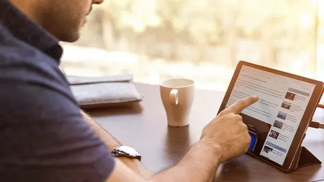 Un homme consulte une tablette numérique sur une table en bois, avec une tasse et un vêtement plié à côté.