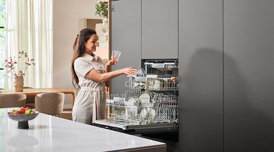 A kitchen setting with a dishwasher loaded with dishes and glassware, set in grey cabinetry. A white countertop shows a bowl of fruit.