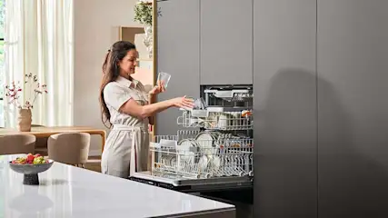 A kitchen setting with a dishwasher loaded with dishes and glassware, set in grey cabinetry. A white countertop shows a bowl of fruit.
