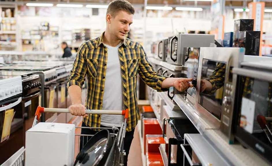 A store aisle displays microwaves. A shopping cart with a white box and pans is to the left.