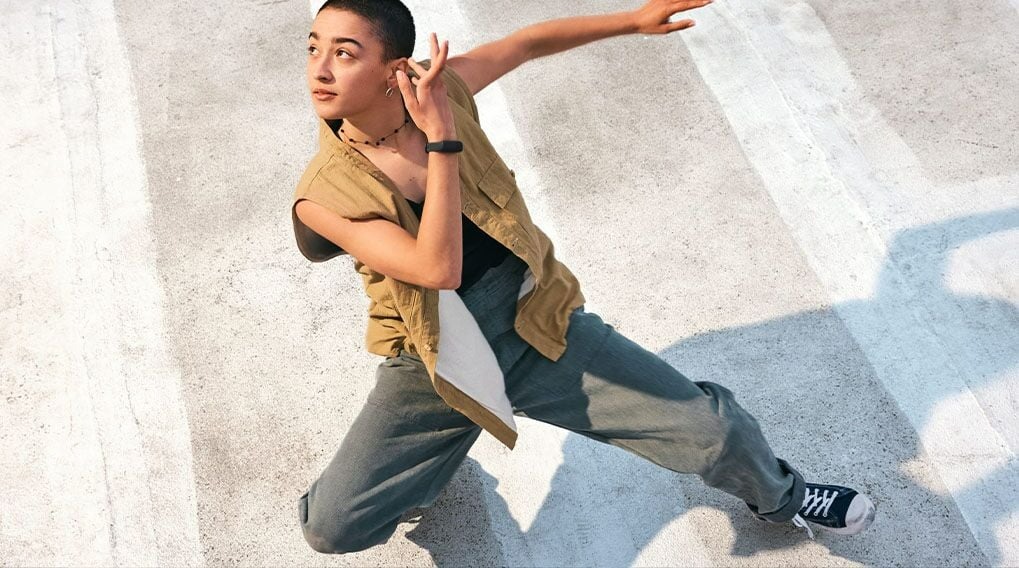 Een persoon met een kaki vest en grijze broek staat in een dansende pose op een betonvloer met witte strepen.