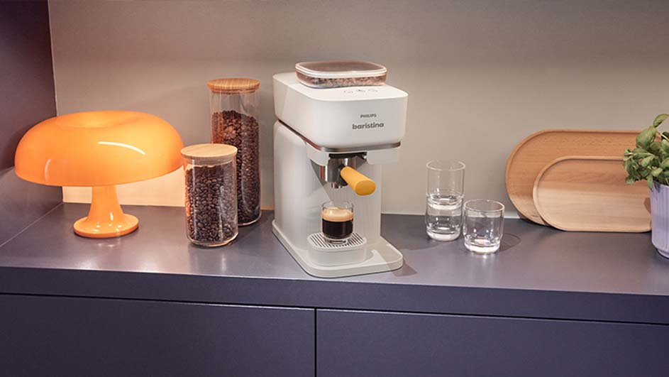 Une machine à café blanche prépare un espresso sur un comptoir gris avec une lampe orange et des bocaux de grains de café.