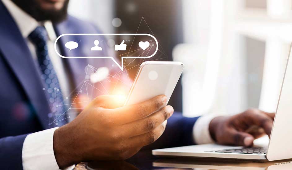 Een hand houdt een witte smartphone met chat-, like- en hartpictogrammen erboven, naast een witte laptop.