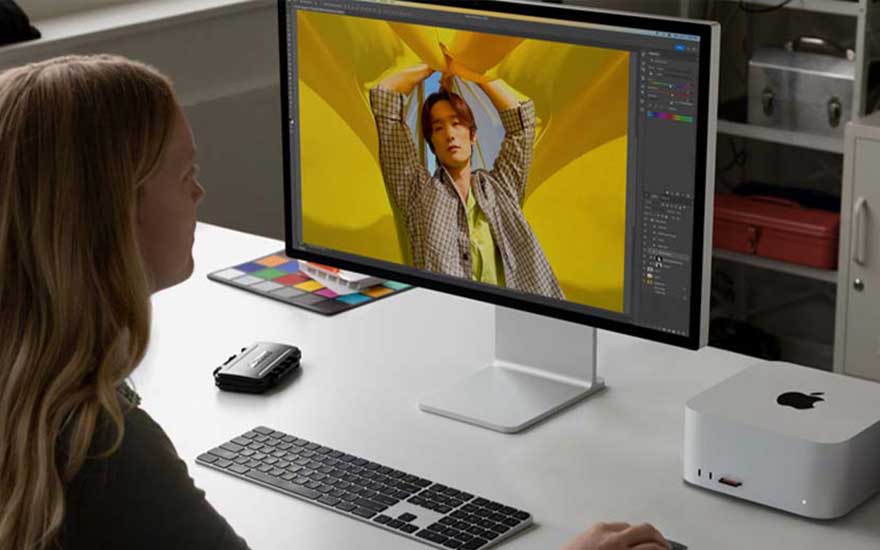 Een bureau met een computerscherm, toetsenbord, muis en een Mac Studio. Op het scherm is een persoon te zien met een gele achtergrond.