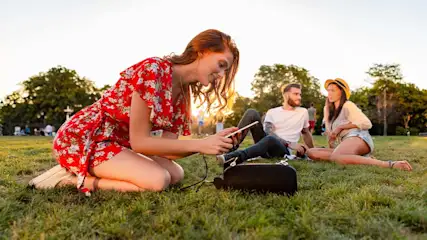 Een groep mensen zit op het groene gras in een park. Een telefoon is aangesloten op een zwarte speaker. Bomen en een heldere hemel zijn zichtbaar op de achtergrond.