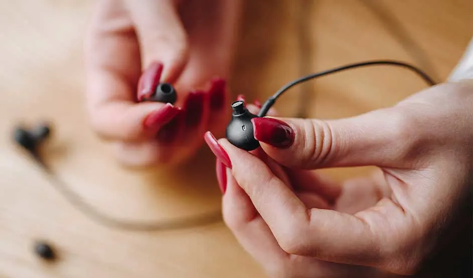 Des mains aux ongles rouge foncé tiennent un écouteur intra noir, l’embout en silicone retiré, devant une surface en bois clair légèrement floue.