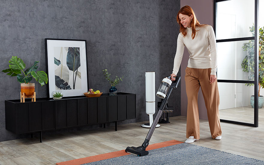 Une femme utilise un aspirateur sans fil sur un tapis dans un salon. Un meuble noir est visible en arrière-plan.