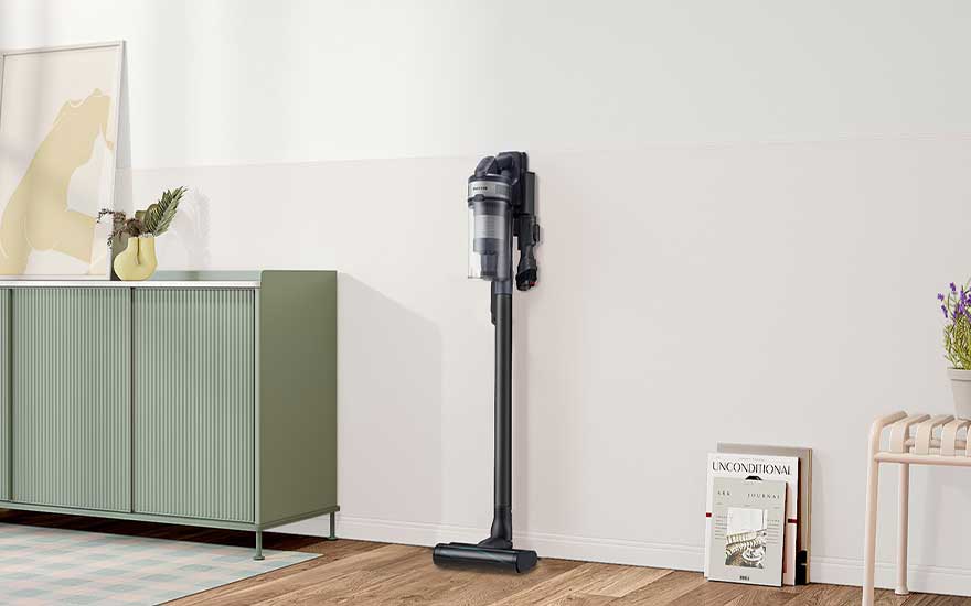 Aspirateur balai gris foncé fixé au mur dans une pièce lumineuse, à côté d’un buffet vert et de magazines posés près du sol.