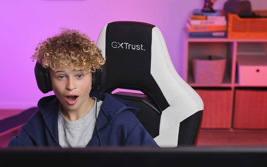 Een zwarte en witte gamestoel staat voor een paarse achtergrond. Een kast met boeken en opbergdozen staat achter de stoel.