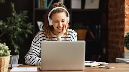 Sur une table en bois se trouvent un ordinateur portable gris, un bloc-notes, un stylo et un téléphone portable. Des plantes et une bibliothèque sont visibles en arrière-plan.