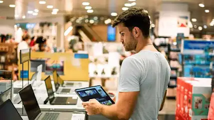 A brightly lit store displays laptops and a tablet being held. Shelves and lights fill the background.