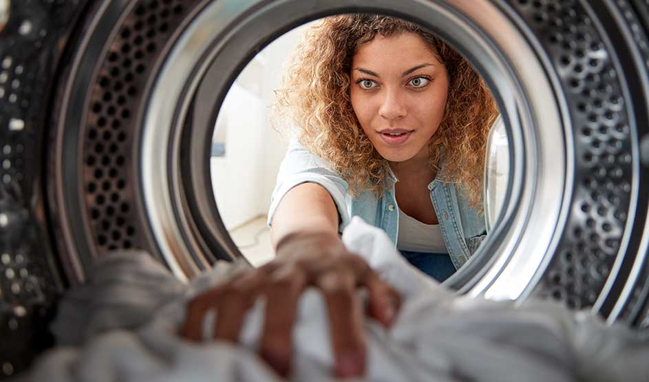 Jonge vrouw kijkt door de wasmachinetrommel en reikt naar witte was; close-up, metalen rand en gaatjes, helder licht, focus op gezicht.
