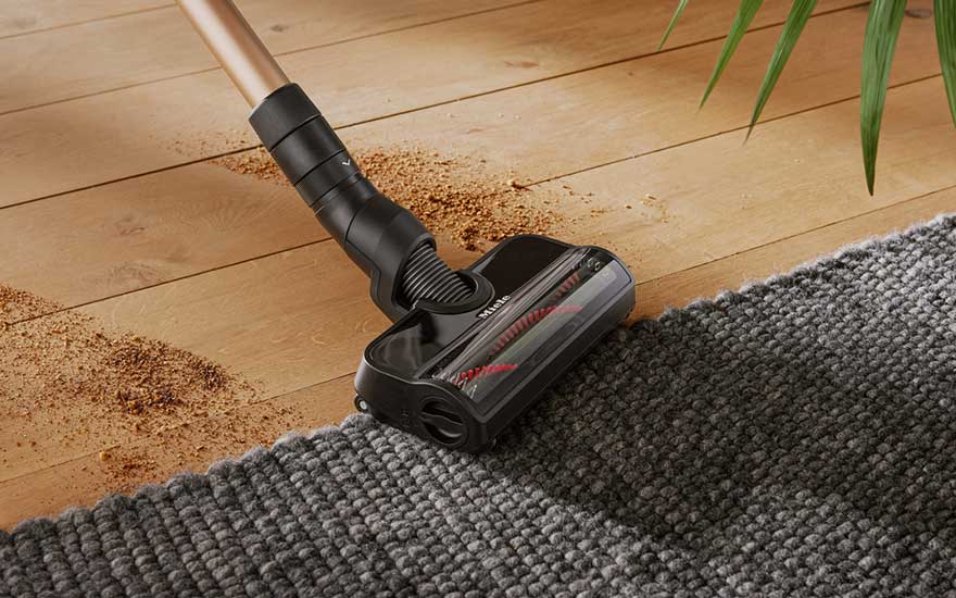 Een stofzuiger met een zwarte kop ligt op een grijze mat en een houten vloer. Er ligt vuil op de vloer.
