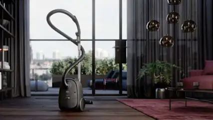 Un aspirateur gris se dresse dans un salon moderne avec des rideaux gris et un grand balcon. Des luminaires suspendus sont visibles.