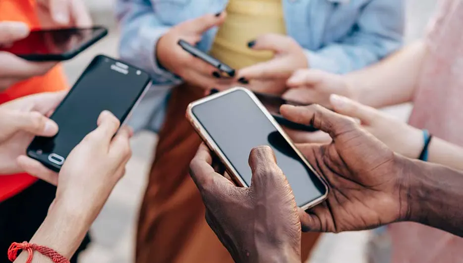 Diverse handen houden smartphones vast in een kring. Schermen zijn zwart, randen goud of zwart. Kleding is kleurrijk, de achtergrond is vaag.
